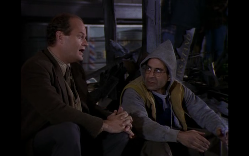 Frasier and Manu sit in front of Manu's burned newspaper stand.