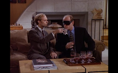 Niles holds a silver spittoon for a blindfolded Frasier to spit his wine into during a tasting practice. 